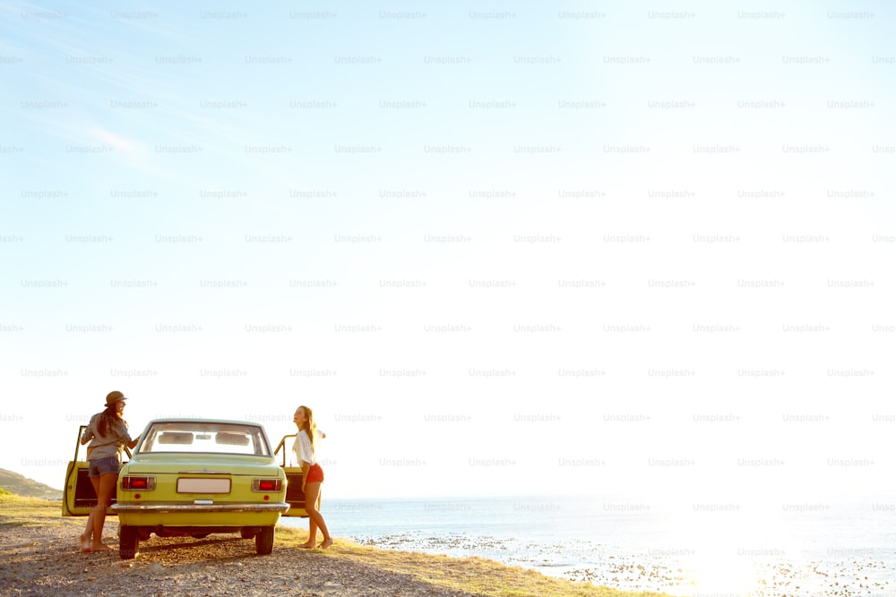 Dos mujeres jóvenes afuera de su automóvil en la playa después de un viaje por carretera