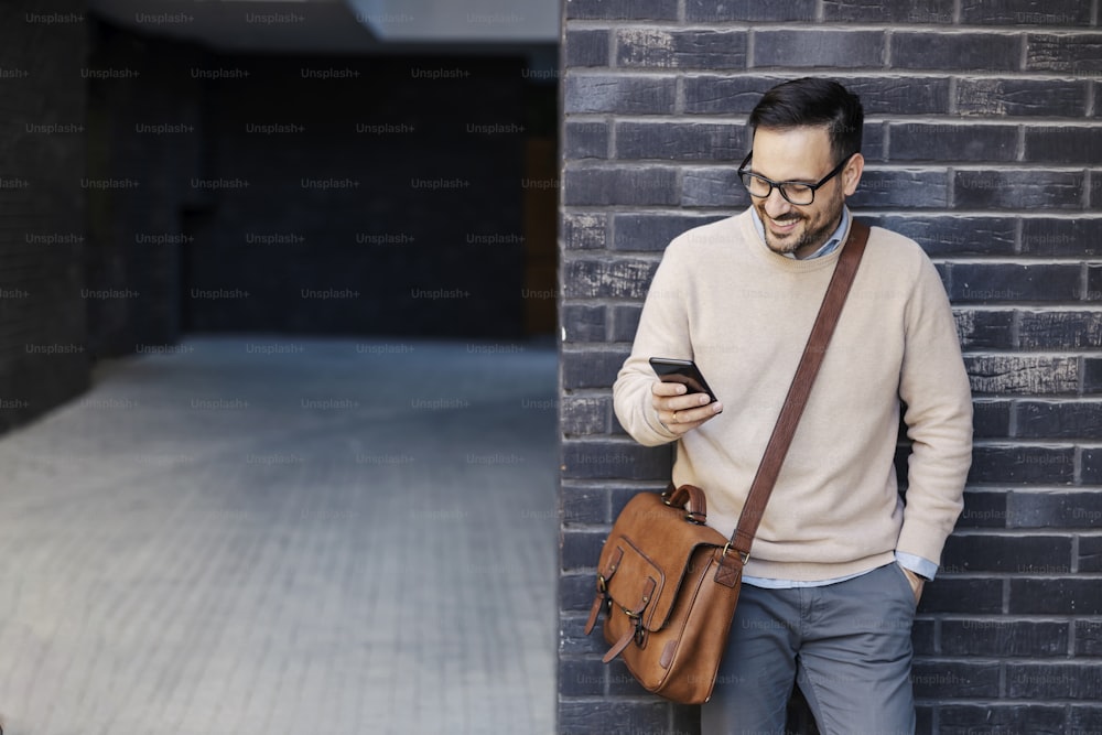 A happy man leaning on the wall on the street, using phone and smiling at it.