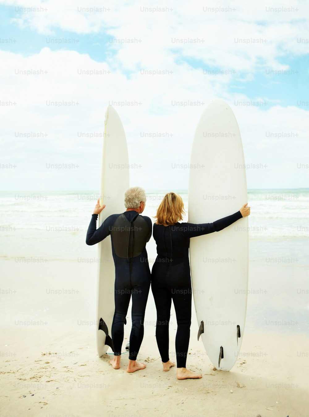 Rearview of an older couple in wetsuits standing with their surfboards