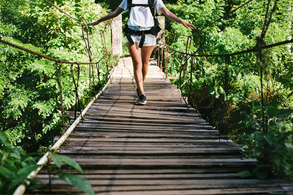 Mulher nova do ajuste com mochila que viaja através da ponte suspensa na floresta tropical