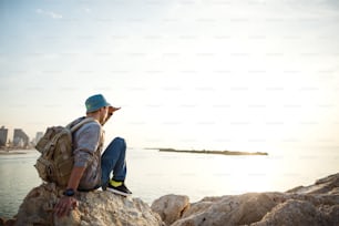 viaggiatore con zaino seduto sugli scogli vicino al mare sulla spiaggia in città e guardando lontano all'orizzonte