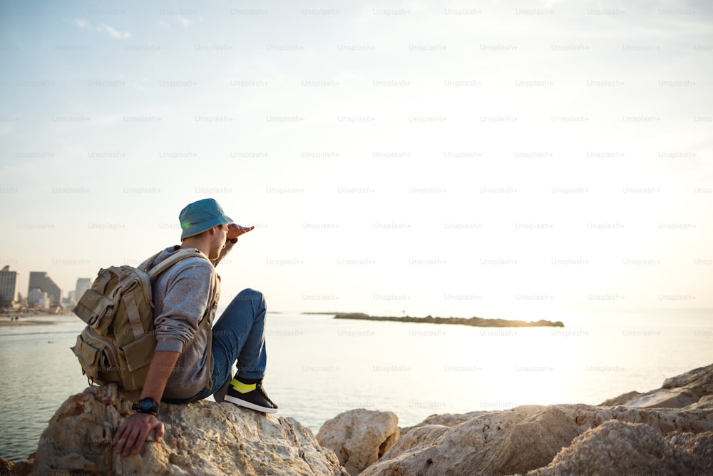 traveler with backpack sitting on the rocks near the sea on the beach in the city and looking far away at the horizon