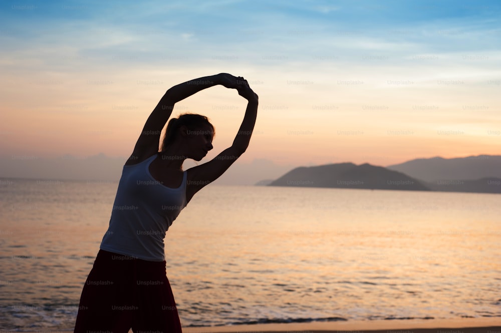 Silhouette de femme sportive active s’étirant et faisant de l’exercice sur la plage au coucher du soleil.