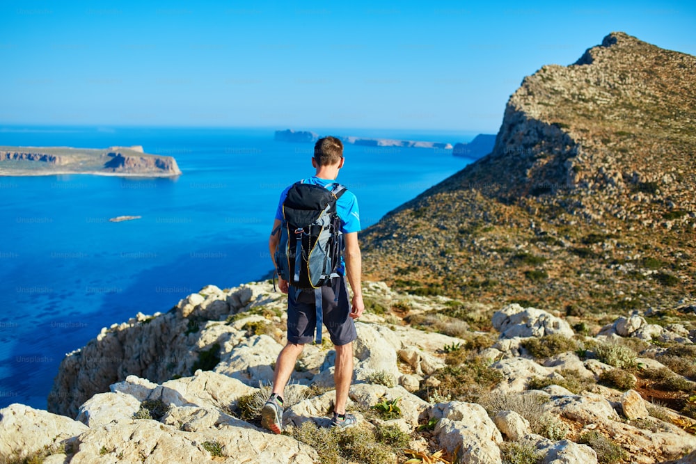 male traveler with backpack runs on the cliff against sea and blue sky at early morning. Balos beach on background, Crete, Greece