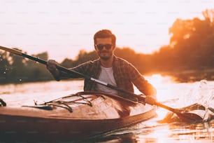 Joven hombre sonriente navegando en kayak en el río con la puesta del sol en el fondo