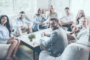 Group of confident young business people sitting around office desk together and looking at camera with smiles