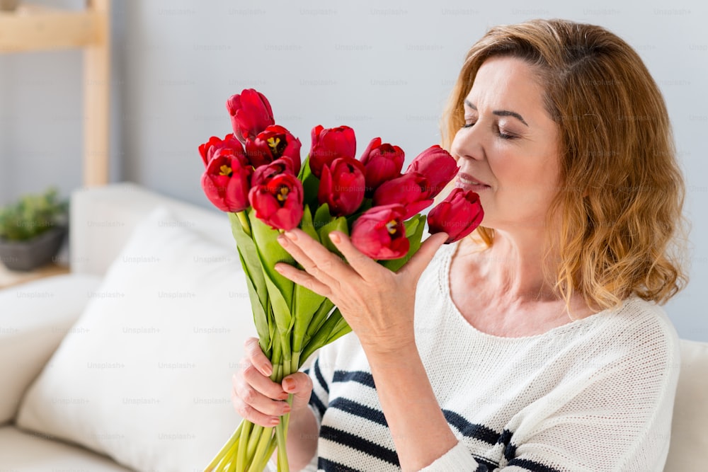 Petty senior woman is holding flowers and smelling it with pleasure. She is smiling happily. Woman is sitting on couch at home