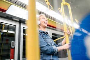 Schöne junge blonde Frau im Jeanshemd steht mit weißen Kopfhörern in der U-Bahn, hört Musik und lacht