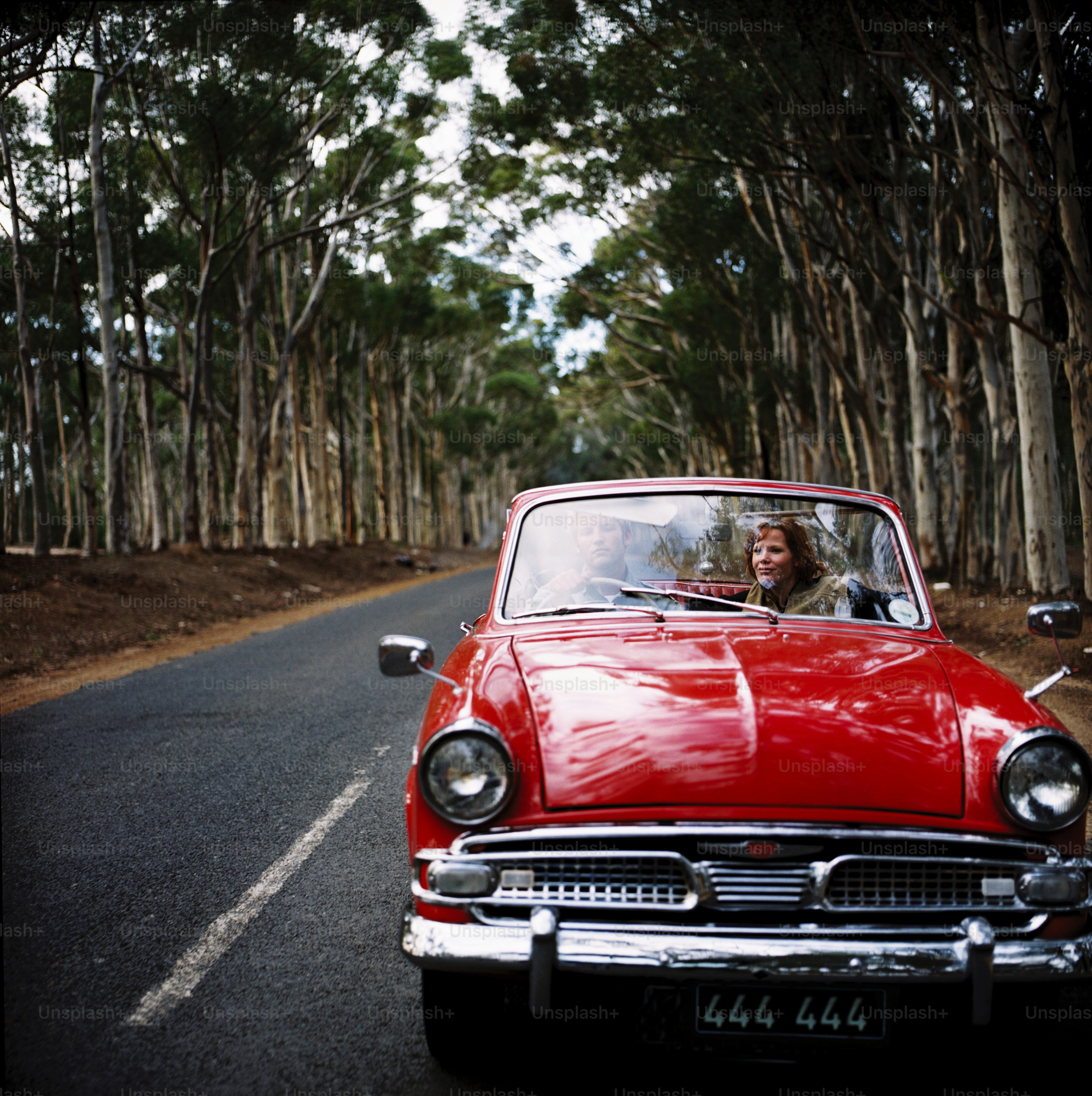 A red car driving down a road next to a forest photo – Vintage car ...