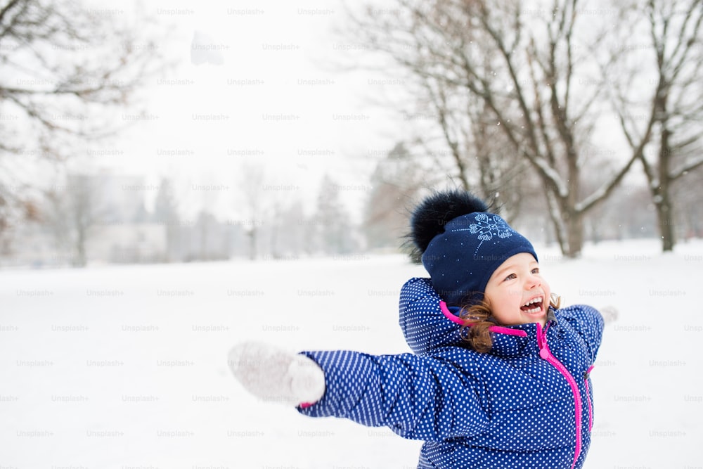 Bambina carina in giacca blu e cappello lavorato a maglia che gioca fuori nella natura invernale, braccia tese