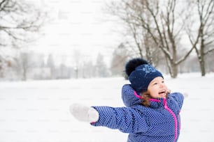 Cute little girl in blue jacket and knitted hat playing outside in winter nature, arms stretched