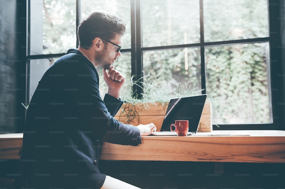 Seitenansicht eines selbstbewussten jungen Mannes mit Brille, der am Laptop arbeitet, während er im Büro oder Café sitzt