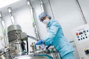 scientist in blue lab uniform stand near big pressure tank , hold tablet , look at camera, turned picture, stand near control panel