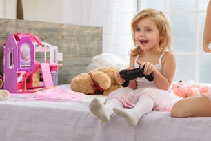 Menina animada está jogando console com sua irmã mais velha. Ela está sentada na cama e rindo