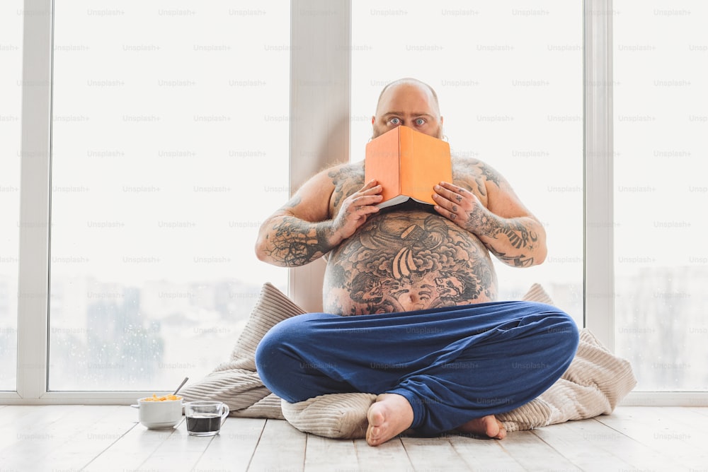 Mature thick guy is scared after reading book. He is hiding behind pages and staring at camera with shock. Fatso is sitting on floor near cup of coffee and cereals
