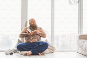 El gordo inteligente está leyendo un libro en casa. Está sentado cerca de la ventana y desayunando