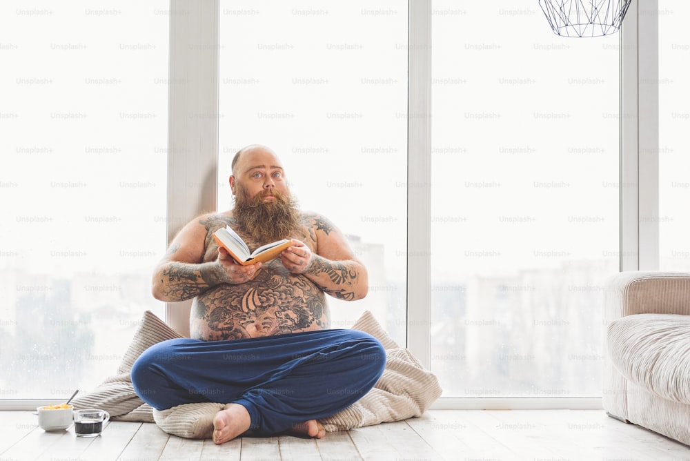 Smart fat man is reading book at home. He is sitting near window and having breakfast