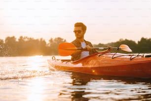 Joven guapo navegando en kayak en el lago con la puesta de sol en el fondo