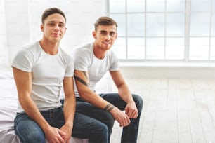 Relaxed young men are sitting on bed and smiling. They are looking at camera with happiness
