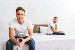 Happy young man is enjoying spending time with his boyfriend. He is looking at camera and smiling. His partner is sitting on bed and holding gadget