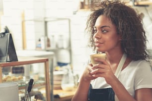 Dreamed woman holding glass of tasty milk coffee while standing near shelf with cakes confectionary shop