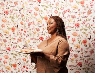 a woman holding a plate of food in front of a floral wallpaper
