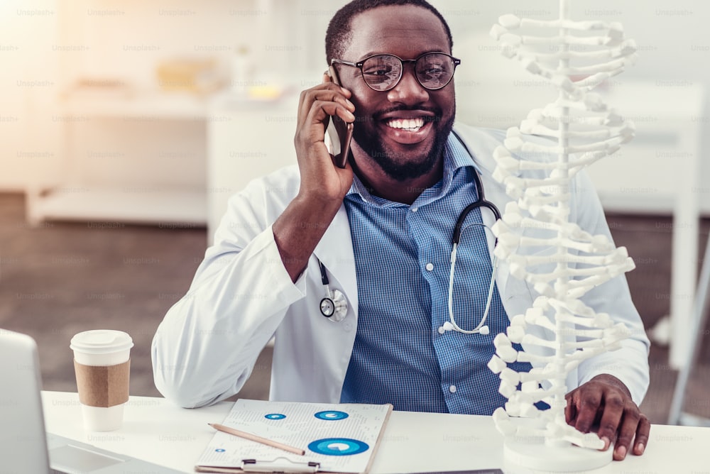 Buenas noticias. Hombre milenario radiante con bata de laboratorio sonriendo alegremente mientras está sentado en su mesa y mirando un modelo tridimensional de ADN durante una agradable conversación telefónica a solas.