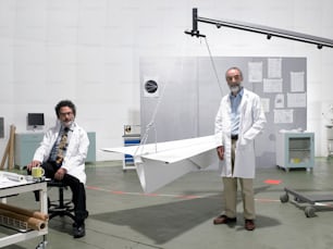 two men in lab coats standing next to a paper airplane