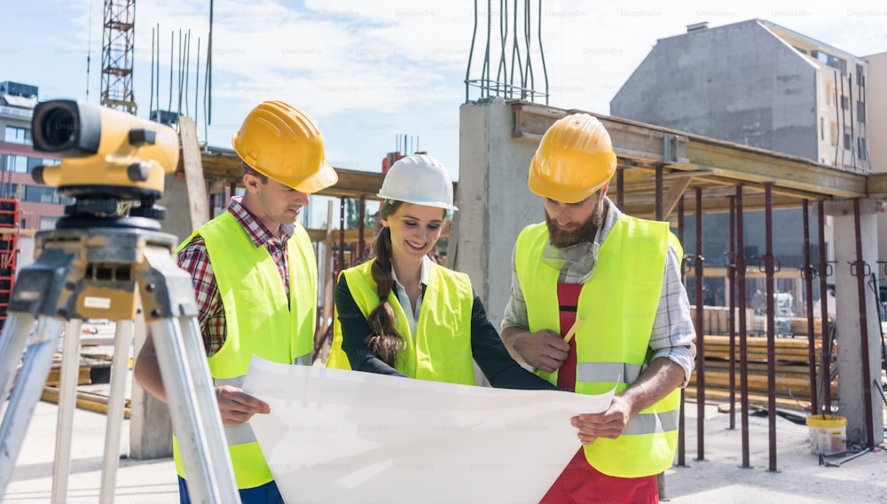 Une architecte expérimentée souriant tout en analysant un plan avec deux travailleurs à l’extérieur sur le chantier de construction d’un immeuble résidentiel moderne