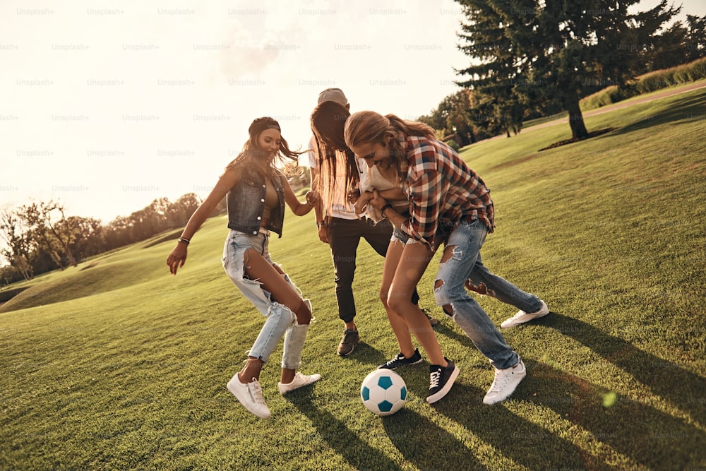 Longitud completa de jóvenes sonrientes en ropa casual disfrutando de un agradable día de verano mientras juegan al fútbol al aire libre
