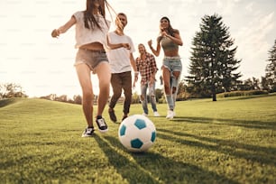 Grupo de jovens sorridentes em trajes casuais correndo enquanto joga futebol ao ar livre