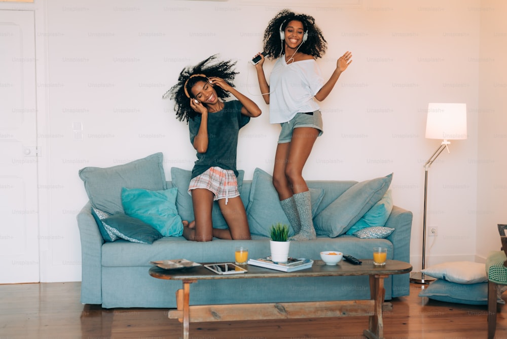 Glückliche junge zwei schwarze Frauen sitzen und stehen auf der Couch auf dem Computer und Handy mit Kopfhörern tanzen "n
