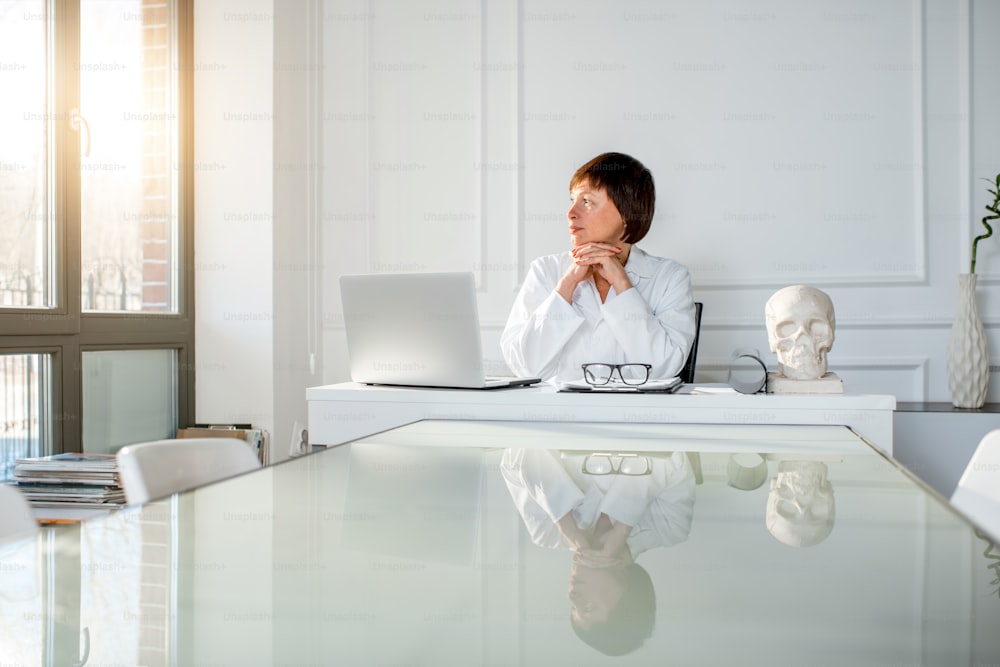 Portrait of a senior woman doctor sitting with laptop in the beautiful white office interior. Wide angle view with reflection on the table