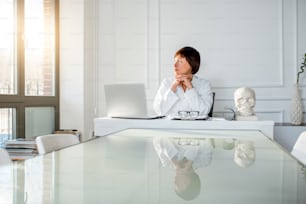 Portrait d’une femme médecin senior assise avec un ordinateur portable dans le bel intérieur blanc du bureau. Vue grand angle avec réflexion sur la table