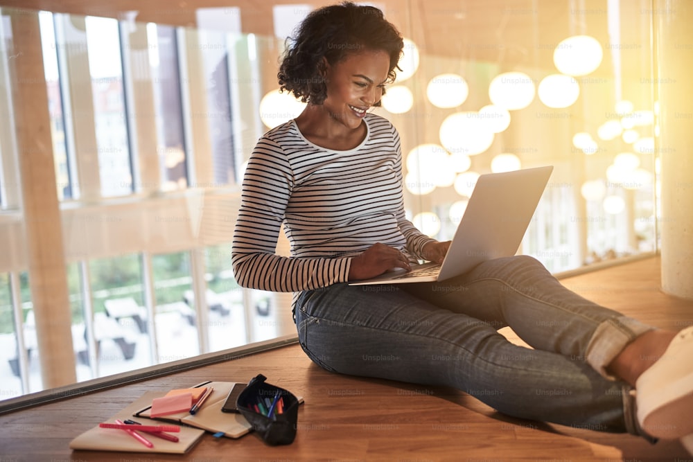 Joven estudiante universitario africano sonriente sentado en el suelo en un pasillo del campus estudiando en línea con una computadora portátil