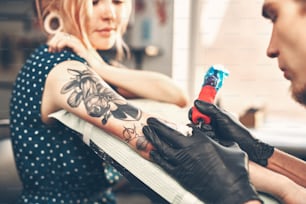 La joven hermosa parece que el tatuador hace el tatuaje, leyendo las noticias en el teléfono inteligente