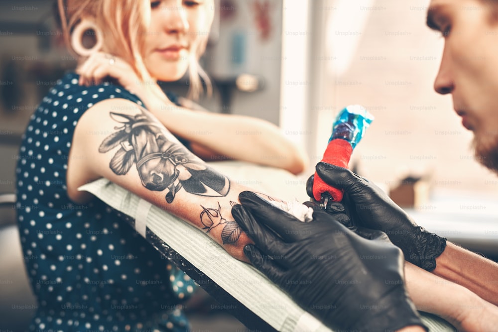 Young beautiful girl looks like the tattoo artist does the tattoo, reading the news in the smartphone
