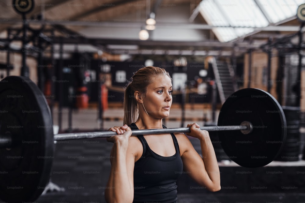 Fitte junge blonde Frau in Trainingskleidung, die sich anstrengt, während sie schwere Gewichte alleine in einem Fitnessstudio stemmt