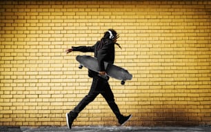 Bonito jovem skatista dreadlocks com fones de ouvido correndo em um terno perto da parede amarela na rua.