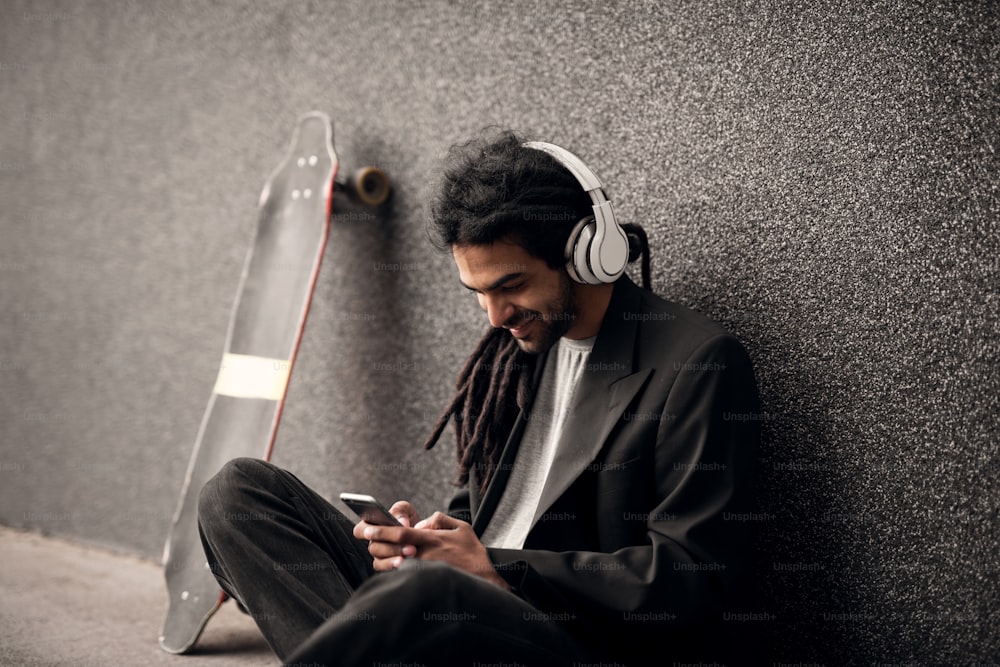 Stylish young dreadlock hipster with headphones sitting leaning against the grey wall and skate near him looking on a mobile.