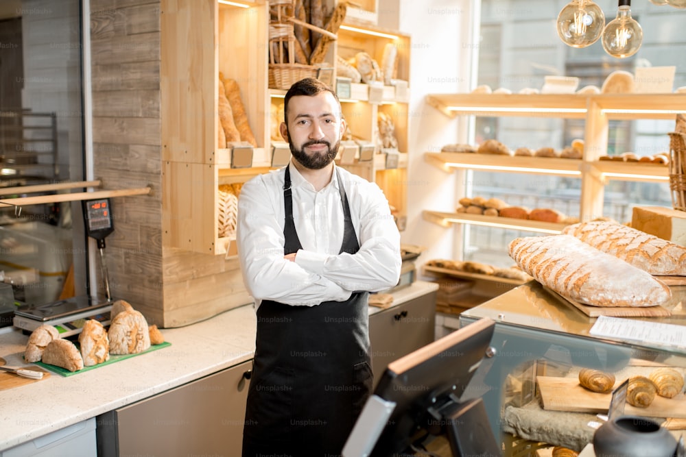 Portrait of a handsome seller in uniform standing in the small and beautiful store with bakery products