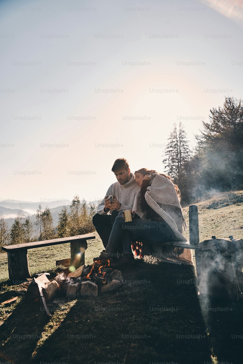 Beau jeune couple prenant le café du matin assis près du feu de camp dans les montagnes