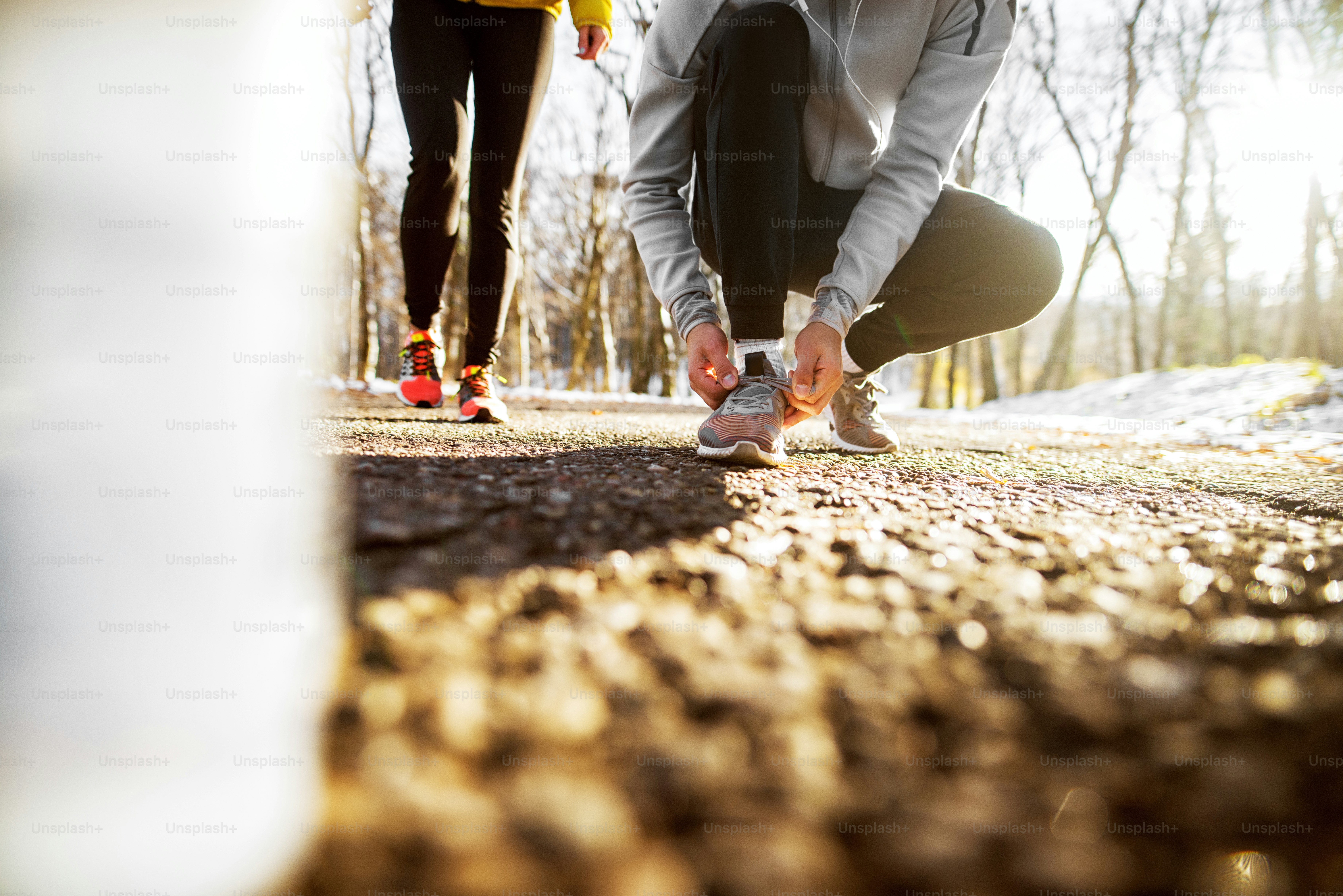 Close up of sport active man in sportswear kneeling on the road and tying shoelaces in the sunny winter morning outside in nature with a girl next to him.