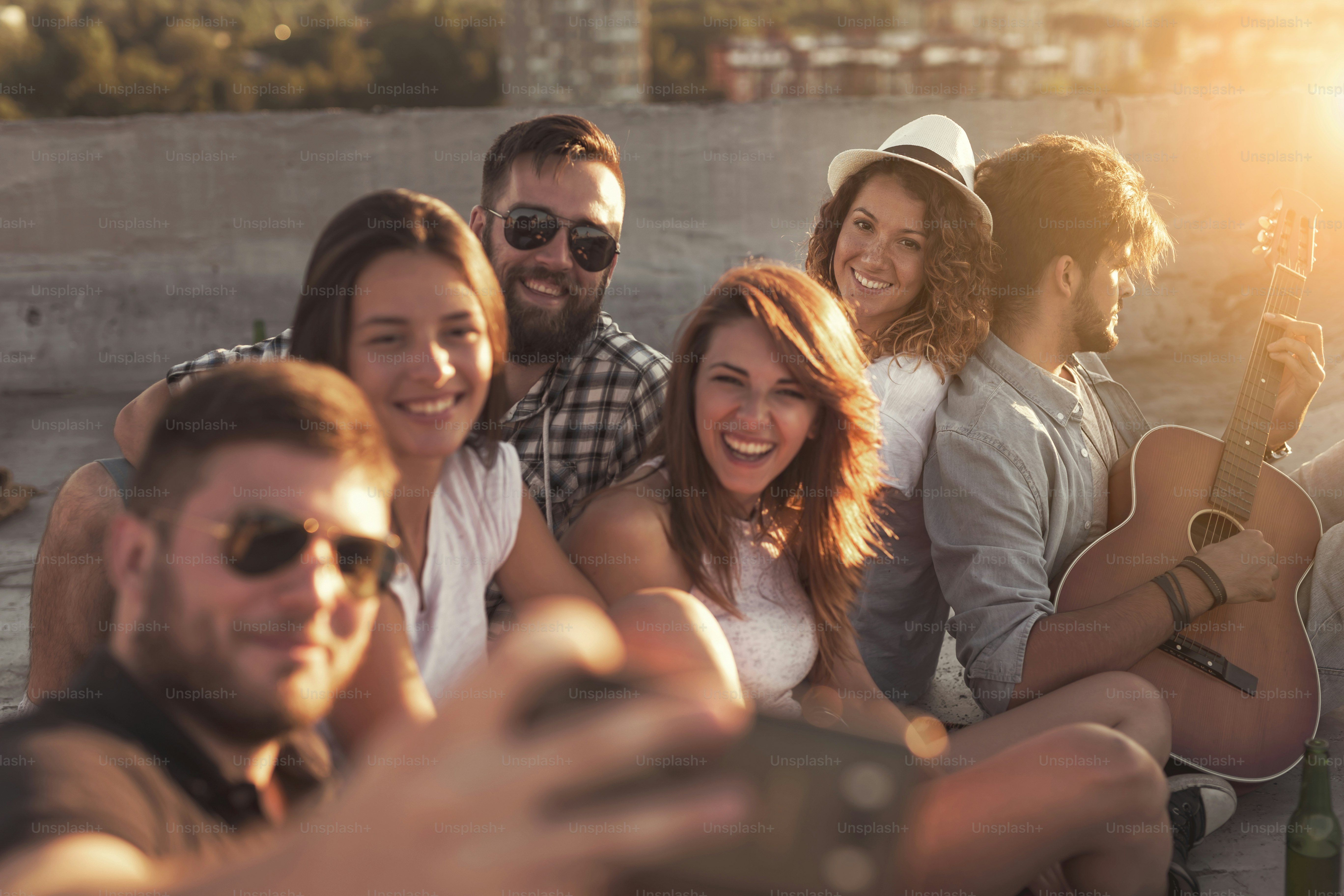Group of young people playing the guitar, singing and taking selfies at a rooftop party. Focus on the girl with a hat