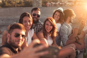 Group of young people playing the guitar, singing and taking selfies at a rooftop party. Focus on the girl with a hat