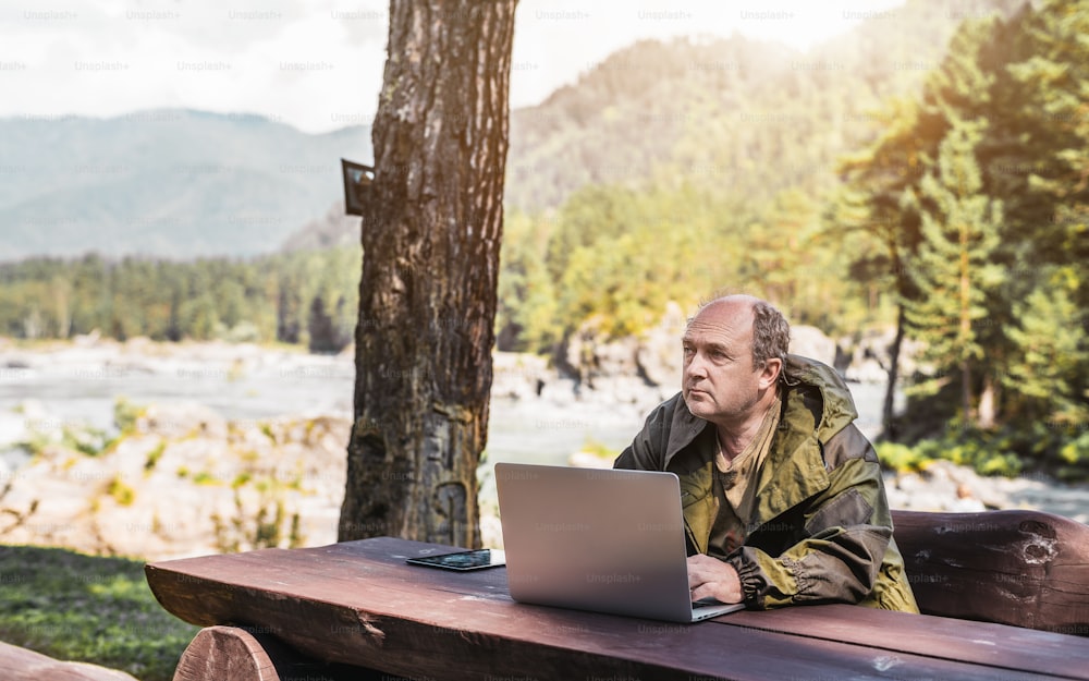 Ein älterer männlicher Jäger sitzt an einem Holztisch im Freien mit Wald und Fluss im Hintergrund und schaut zur Seite, während er sein Netbook benutzt. mit digitalem Tablet in der Nähe und Kopierplatz für Werbung