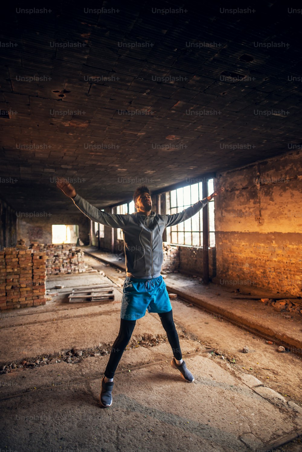 Portrait of active afro-american young attractive athletic man with earphones stretching and warming up before workout inside of the abandoned place with open arms.