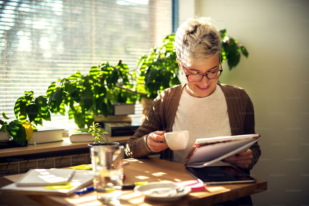 Charmante, fröhliche, zufriedene Frau mit kurzen Haaren mittleren Alters, die morgens zu Hause am Tisch sitzt und einen Kaffee trinkt, während sie Schülertests oder Unternehmensstatistiken liest.