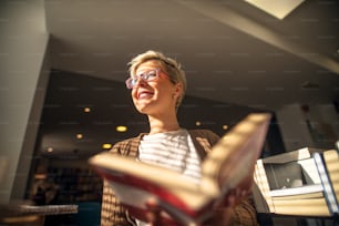 Close up of happy beautiful satisfied middle age short hair woman with eyeglasses sitting on the sofa in the library and looking through the sunny window while holding a book.