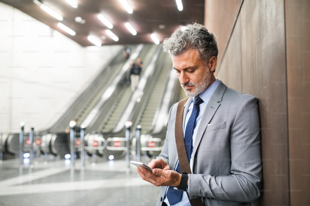 Handsome mature businessman in a city. Man with smartphone standing in front of an escalators at the subway station, text messaging.